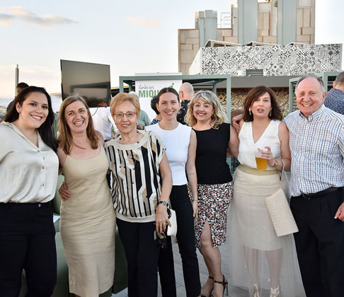 Siete personas posando juntas en una terraza al atardecer, con una pantalla y un cartel al fondo